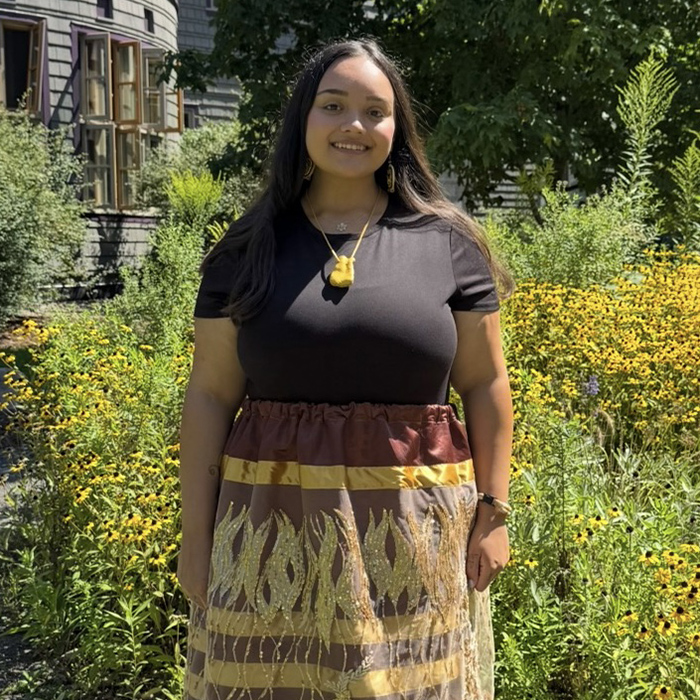 Young woman with long brown hair and a brown shirt and colorful long skirt