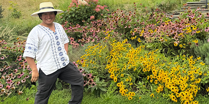 young man with a brimmed hat, brown hair and white shirt stnading next to a hillside of colorful flowers