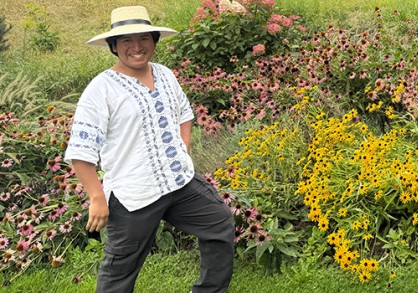young man with a brimmed hat, brown hair and white shirt stnading next to a hillside of colorful flowers