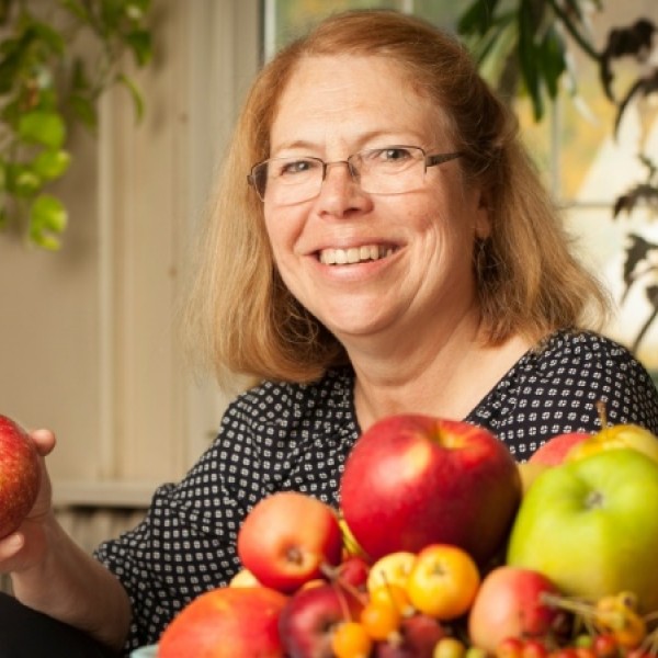 A smiling woman holds an apple next to more apples piled on a table