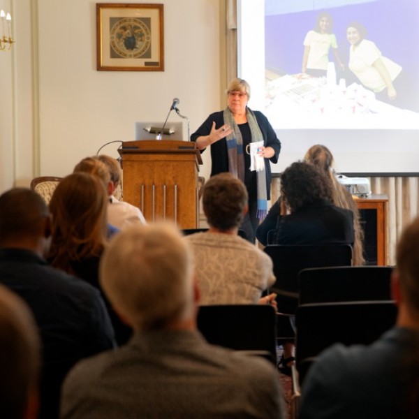 An older white woman standing at the front of the room in front of a screen, leaning on a podium talking to a room full of sitting people.