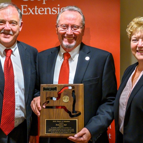 Two white older men and a white older woman standing next to one another smiling for the camera. The middle man is holding a wooden plaque 