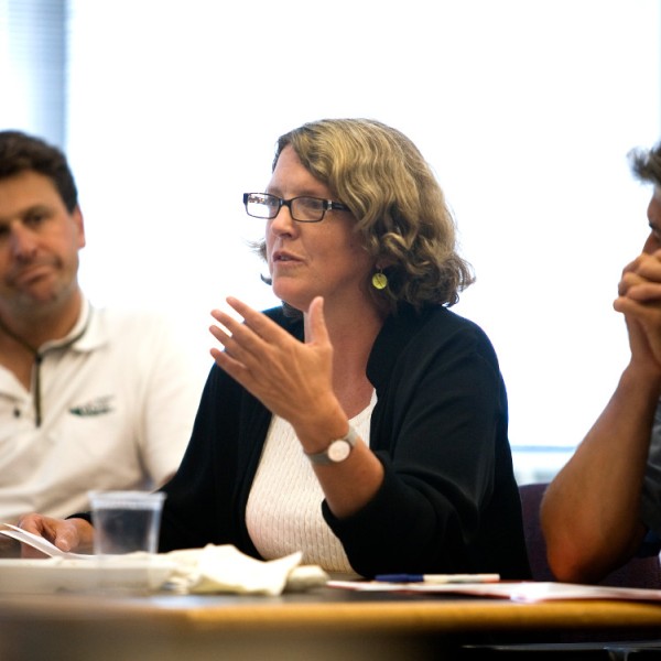 An older woman with grey hair sitting at a table with other adults, speaking with them and gesturing.