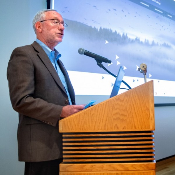 An older, white adult male standing at a podium in front of a screen and speaking to an audience while the rest of the panel looks on.
