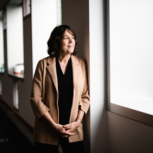 A caucasian woman with brown hair wearing a black shirt and tan jacket standing and looking out a window with her hands clasped in front of her