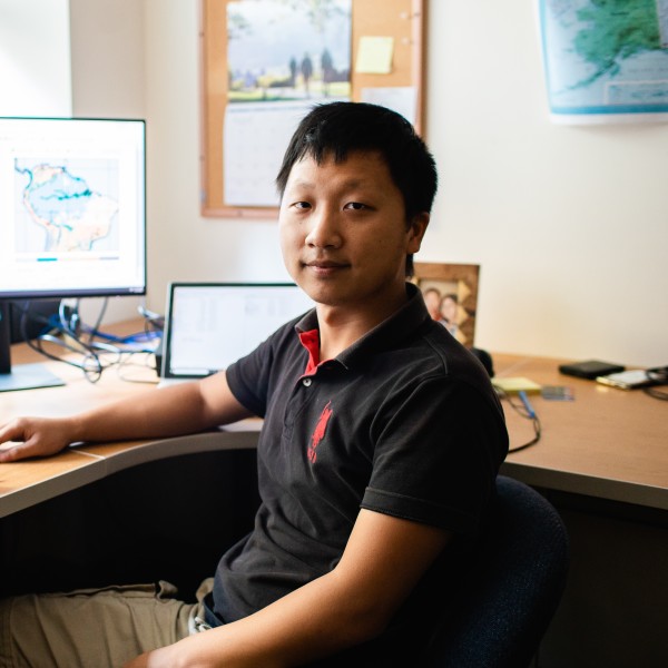 An asian male sitting at a desk in his office with a computer behind him