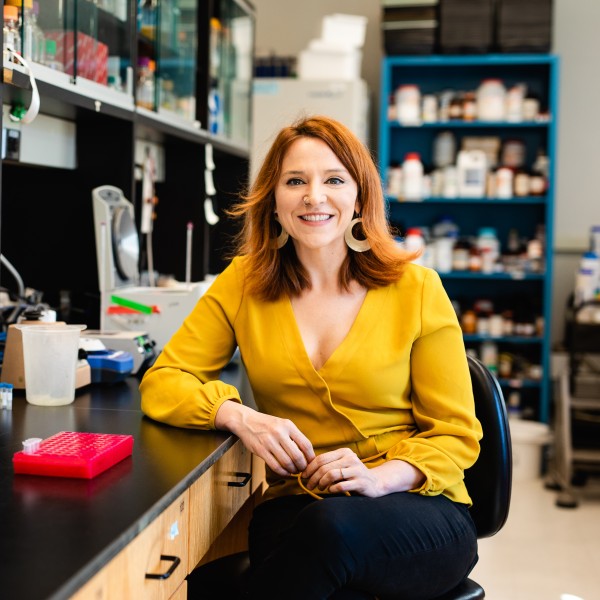 A Caucasian female with red hair in a yellow shirt sitting at a lab bench 