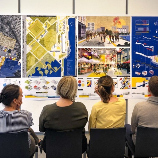 Several men and women sitting with their back to the camera looking at colorful posters of landscape architecture with a young woman in the front of the room presenting the posters
