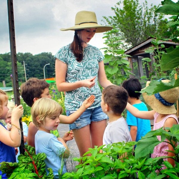 A woman standing in a garden in shorts and a short sleeve shirt and sun hat, surrounded by children looking up to her 