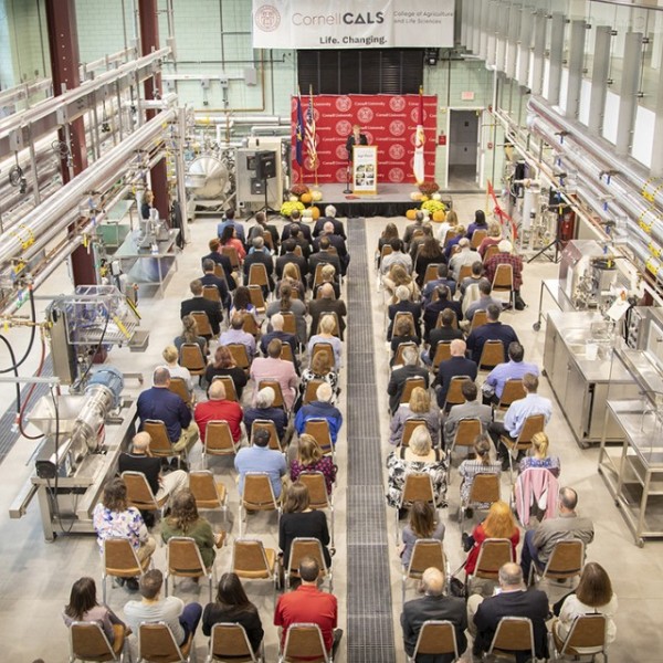 Over fifty people sitting in chairs in a factory-type room all facing ahead listening to a speaker at the front of the room