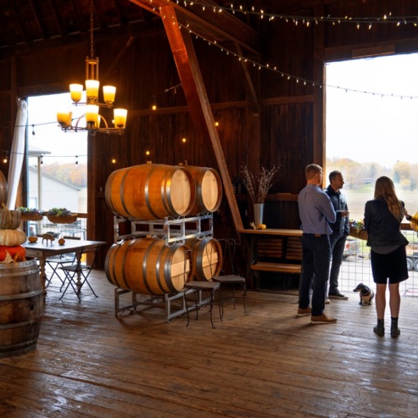 A group of adults standing and talking in a wooden barn containing wine barrel decor