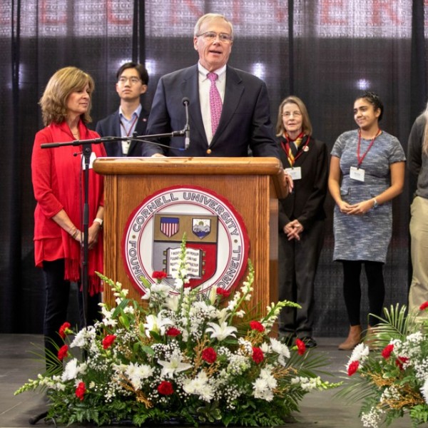 Men and women standing on a stage behind a man standing and speaking at a podium