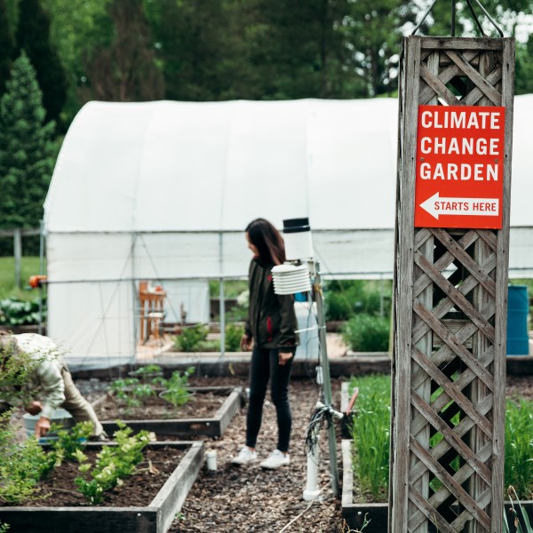 In the foreground, a red sign reads Climate Change Garden. In the background, two women tend to leafy plants in a garden bed.