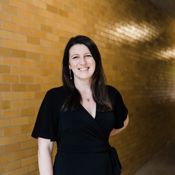 A female with long brown hair in a black dress standing with a yellow brick wall behind her