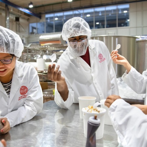 Male and female students in white lab jackets and hair nets standing around a metal table testing ice cream