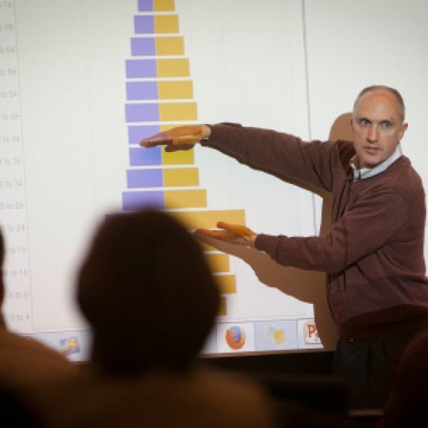 A man lectures in front of a class while pointing to the projection screen