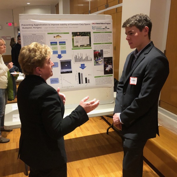 A woman talks to a young man standing in front of a poster that he made