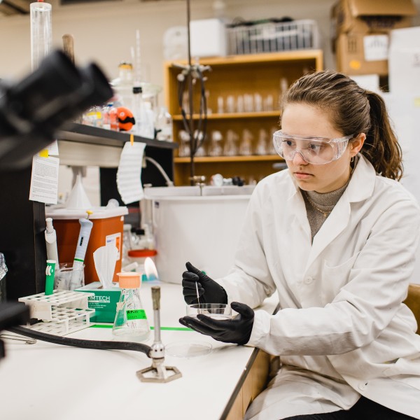 A young woman in a lab coat and safety goggles holds a thin wire near an open flame on a bunson burner