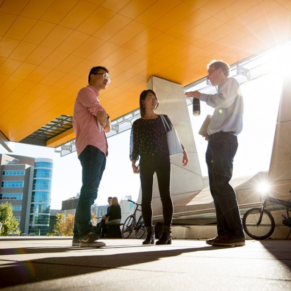 A woman and two men standing outside under a building overhang and talking