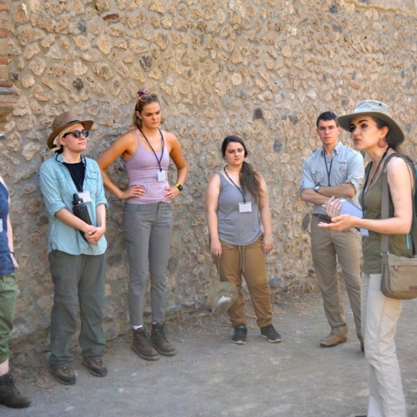Group of five listens to woman speaking as they stand in front of wall