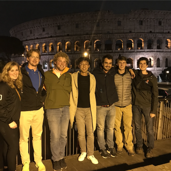A group of seven people stand happily in front of the roman coliseum 