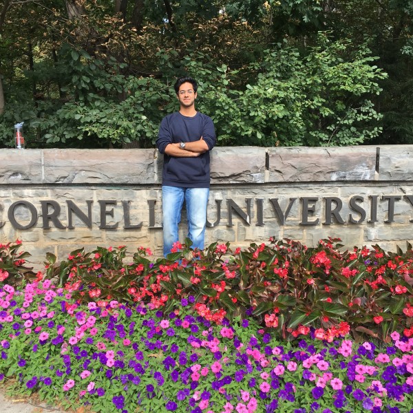 a college-aged man stands in a flower garden by a sign that reads Cornell University