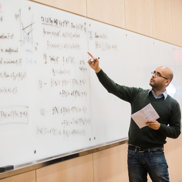 a bald man with glasses writes a complex equation on a whiteboard