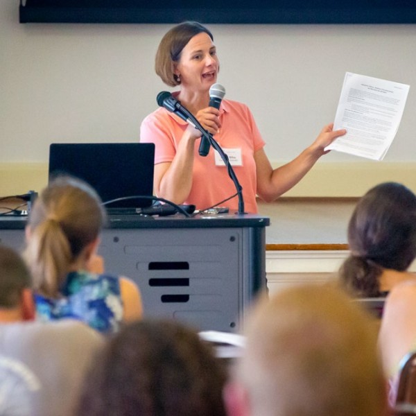 A woman standing at the front of a room of people talking into a microphone 