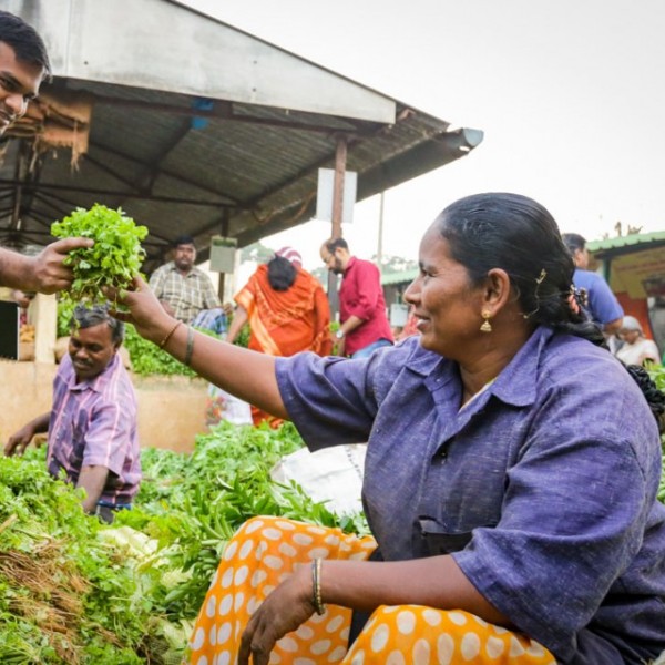 A man standing and taking leafy, green produce from a woman sitting in a market