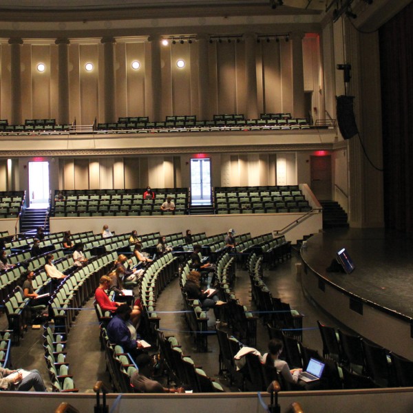 A female faculty member standing on a stage in a large performance hall to students scattered in the chairs below