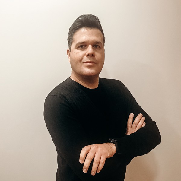 Elad Tako standing in a black sweater with his arms folded against a white wall 