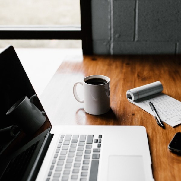 A laptop on a wooden table with a notepad and pen and a cup of coffee