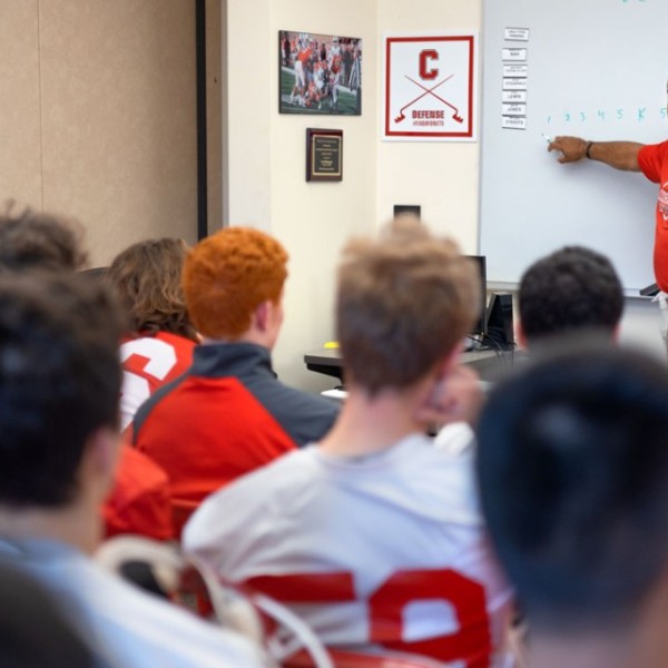 Michael L. Huyghue ’84 stands in a room at a white board and goes over plays with the sprint football team