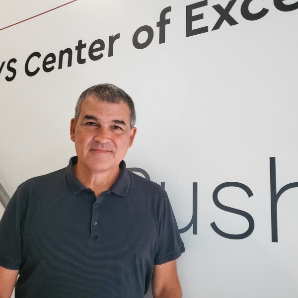 Headshot of Mark Scoville. Man standing on stairs with white background, wearing short-sleeved black polo shirt. 