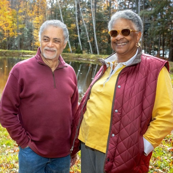 Evon and Sydney Antonio in red fall attire, standing next to a lake, surrounded by trees. Fallen yellow leaves cover the ground. 