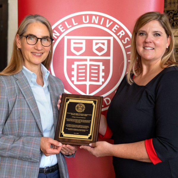 Susan Riley and Caroline Williams pictured in front of a large red banner with the Cornell seal, holding the plaque for the Cornell New York State Hometown Alumni Award.
