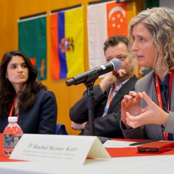 Rachel Bezner Kerr speaks into a microphone, aside other panelists, in front of a wide array of country flags.  