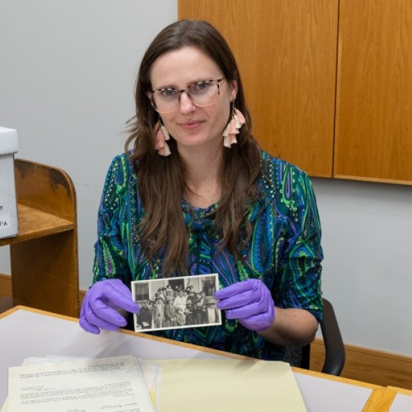 Lynda Xepoleas in an archival room holds up a historical photo of a group of people from her research.