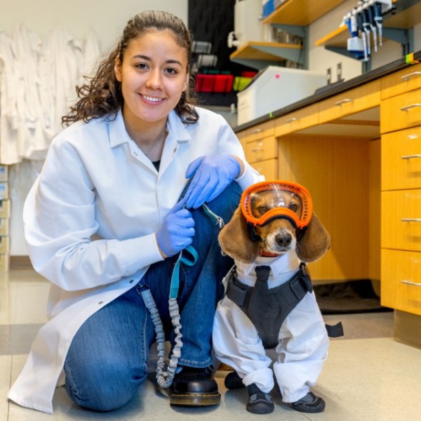 Genesis Contreras and her service dog inside a lab. Genesis is wearing a lab coat, and holding the leash to her dog. The dog is wearing a lab coat, booties, and googles as well.