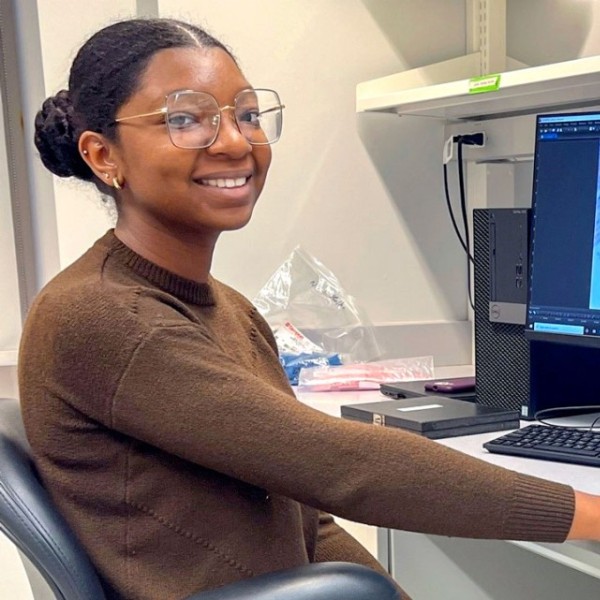 Female student smiles on the left, next to a computer showing an image from a microscope, which is on the far right of the image.