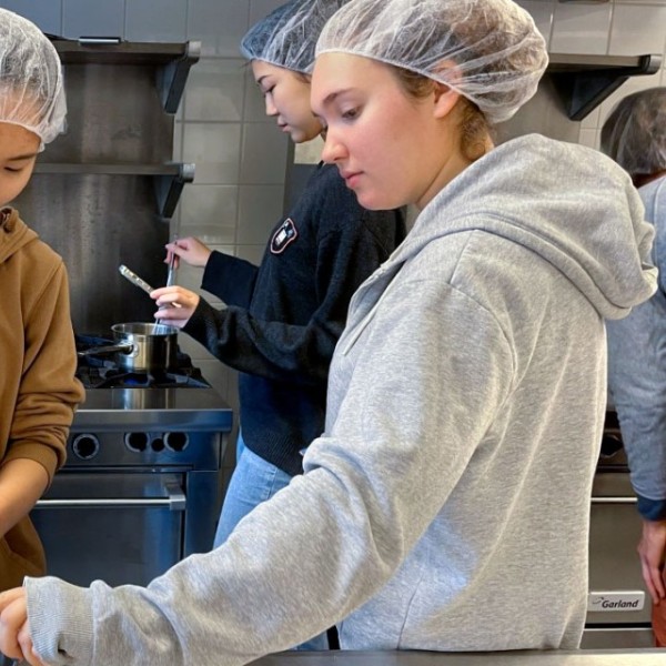 Three women in a kitchen wearing hairnets. Robin Kim on the left is measuring corn syrup, while Jordan Roth holds the measuring cup. Sue Kim in the background takes the temperature of a sugar water solution in a pot on a stove.