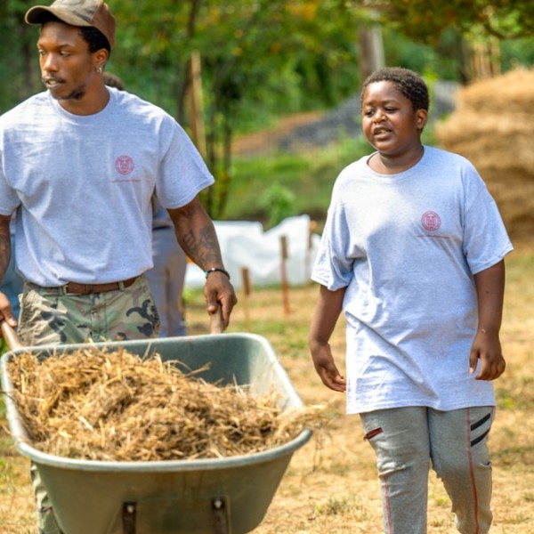 Two young adults in a garden - on pushing a wheel barrow and the other walking alongside him.