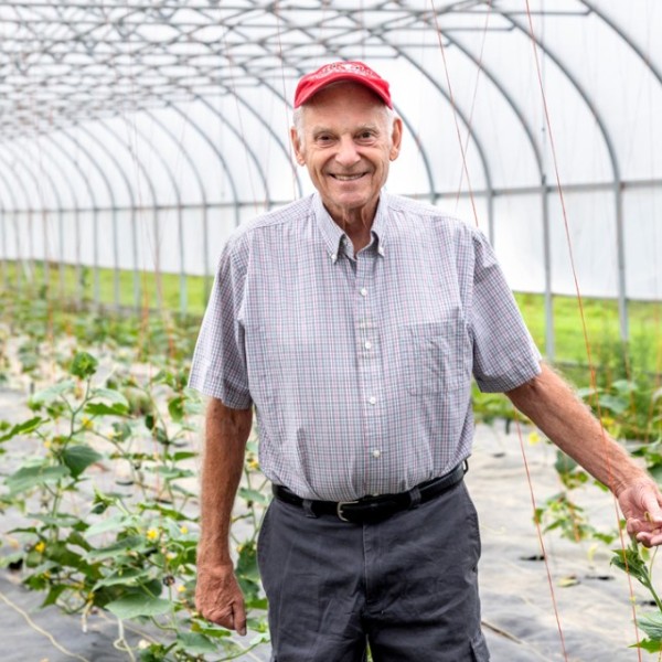 An older man stands in the middle of a greenhouse in a dress shirt and red hat.