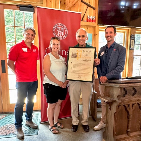 Three men and one woman stand in front of a red Cornell CALS banner with a New York State Assembly Proclamation framed poster.