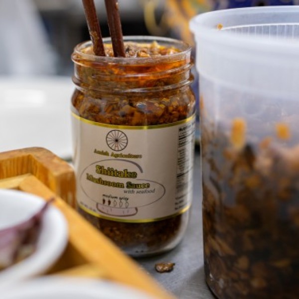 A jar of Amish shiitake mushroom sauce on a table with a plate and other food packaging surrounding it.