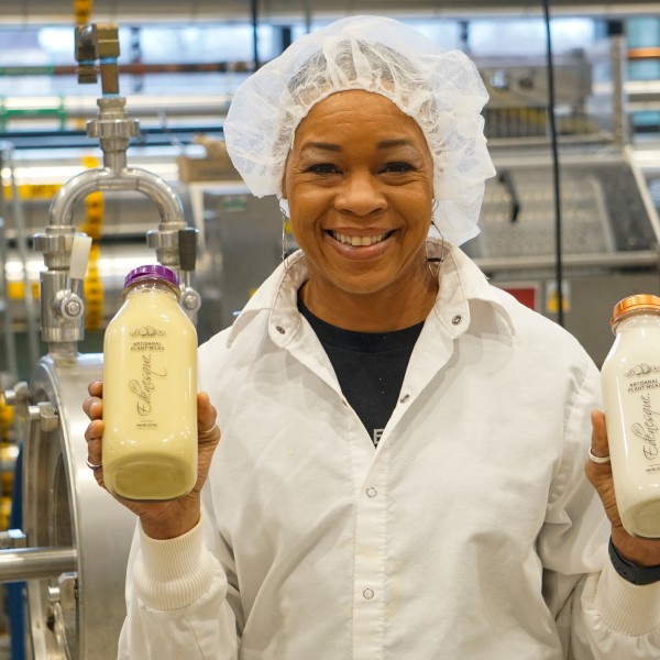 A woman wearing a white lab coat and hair net holding two bottles of milk. 