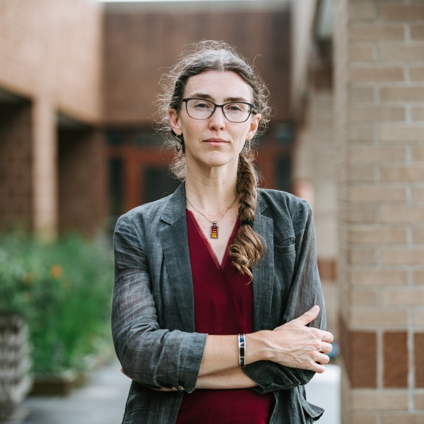 Maria C. Taylor wearing a grey jacket and red shirt, stands in front of a brick building.