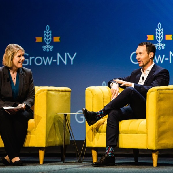 Catharine Young and Benjamin Z. Houlton sit in bright yellow arm chairs talking with "grow-ny" all over the back wall. 