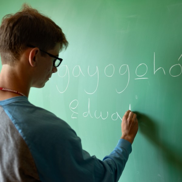 A male student writes Gayogohó:nǫˀ on a green chalk board.