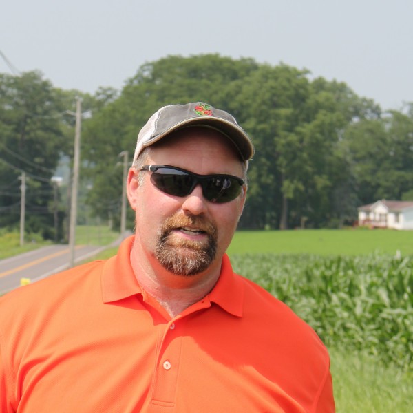 Photo of Andy Miller in an orange shirt and sunglasses, smiling with the farm in the background.